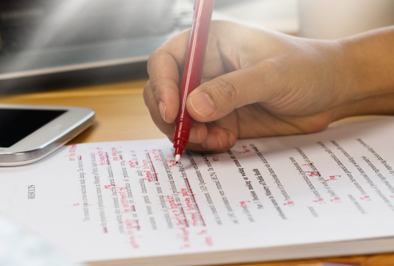 Hand holding red pen over proofreading text in office